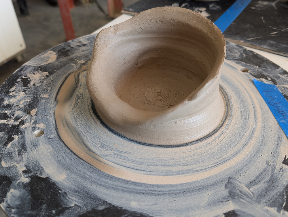 A clay pot on a spinning wheel, with one side collapsed.